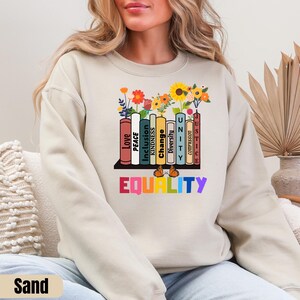 May include: A sand-colored sweatshirt with a graphic of a bookshelf with books that have words like "Love", "Peace", "Inclusion", "Change", "Kindness", "Diversity", "Unity", "Justice", and "Compassion". The words are stacked on top of each other and are in different colors. The graphic is surrounded by flowers and butterflies. The words "EQUALITY" are written in rainbow colors below the bookshelf.