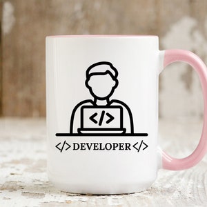 May include: White coffee mug with a pink interior and handle. The mug features a black line drawing of a person working on a laptop, with the word "DEVELOPER" below. The design is simple and modern.