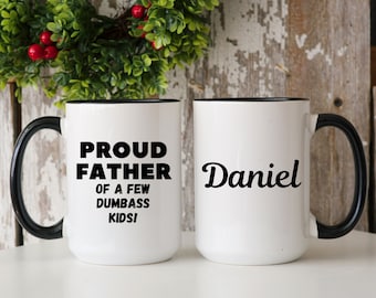 May include: Two white ceramic mugs with black handles and rims. One mug reads "PROUD FATHER OF A FEW DUMBASS KIDS!" in black text. The other mug has the name "Daniel" in black script.