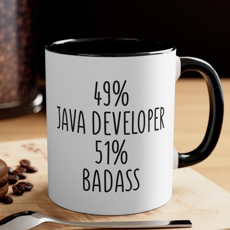 May include: White ceramic mug with a black interior and handle. The mug features the text "49% JAVA DEVELOPER 51% BADASS" in black, bold font. The mug is sitting on a wooden surface.