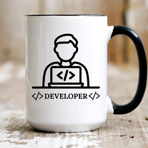 May include: White ceramic mug with a black handle and rim. The mug features a black line drawing of a person working on a laptop, with the word "DEVELOPER" and code symbols. The mug is on a wooden surface.