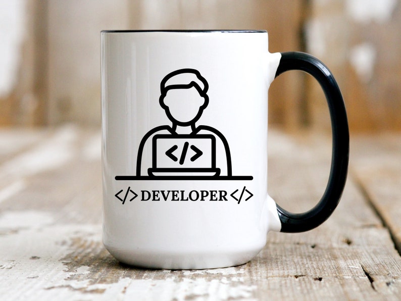 May include: White ceramic mug with a black handle and rim. The mug features a black line drawing of a person working on a laptop, with the word "DEVELOPER" and code symbols. The mug is on a wooden surface.