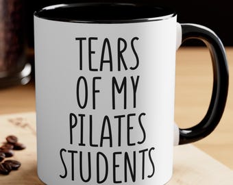 May include: White ceramic mug with a black interior and handle. The mug features the text "TEARS OF MY PILATES STUDENTS" in bold, black letters. The mug is on a wooden surface.