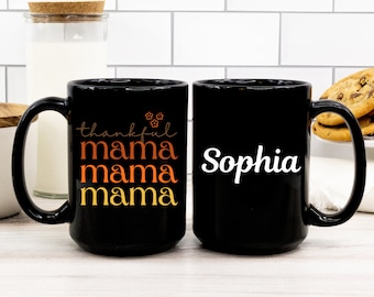 Może przedstawiać: Dwa czarne ceramiczne kubki. Jeden kubek zawiera słowa "thankful mama mama mama" w pomarańczowym i żółtym skrypcie. Drugi kubek ma imię "Sophia" w białym skrypcie. Oba kubki mają czarny uchwyt.