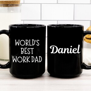 May include: Two black coffee mugs. One mug says "World's Best Work Dad" and the other mug says "Daniel".