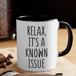 May include: White ceramic mug with a black interior and handle. The mug features the text "RELAX, IT'S A KNOWN ISSUE" in black, bold font. The mug is likely used for coffee, tea, or other beverages.