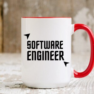 Puede incluir: Taza de cer&aacute;mica blanca con interior y asa rojos. La taza presenta el texto "SOFTWARE ENGINEER" en letras negras en negrita, con detalles de flechas. Sobre una superficie de madera blanca desgastada.