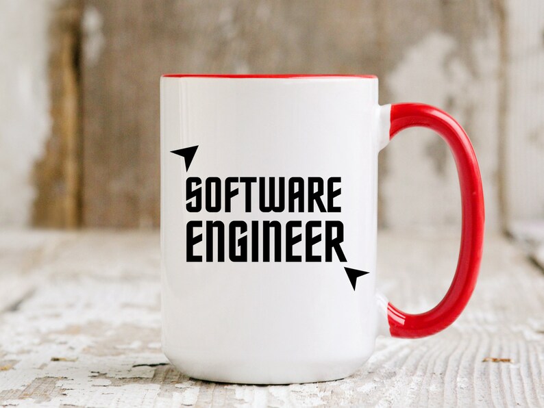Puede incluir: Taza de cer&aacute;mica blanca con interior y asa rojos. La taza presenta el texto "SOFTWARE ENGINEER" en letras negras en negrita, con detalles de flechas. Sobre una superficie de madera blanca desgastada.
