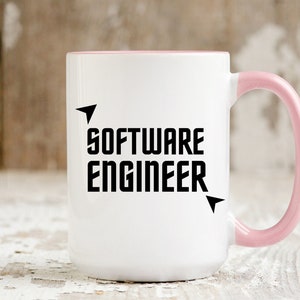 Puede incluir: Taza de cer&aacute;mica blanca con interior y asa rosas. La taza presenta el texto "SOFTWARE ENGINEER" en letras negras en negrita con detalles de flechas. La taza se utiliza probablemente para bebidas.