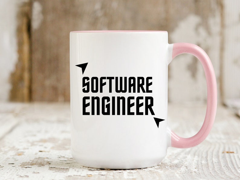 Puede incluir: Taza de cer&aacute;mica blanca con interior y asa rosas. La taza presenta el texto "SOFTWARE ENGINEER" en letras negras en negrita con detalles de flechas. La taza se utiliza probablemente para bebidas.