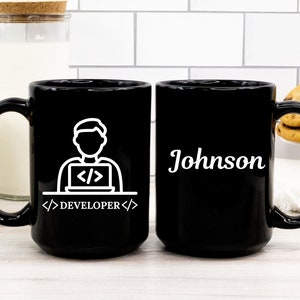 May include: Black ceramic mug with a white graphic of a person working on a laptop, with the word "DEVELOPER" below. The other side of the mug has the name "Johnson" in white. The mug is on a white surface.