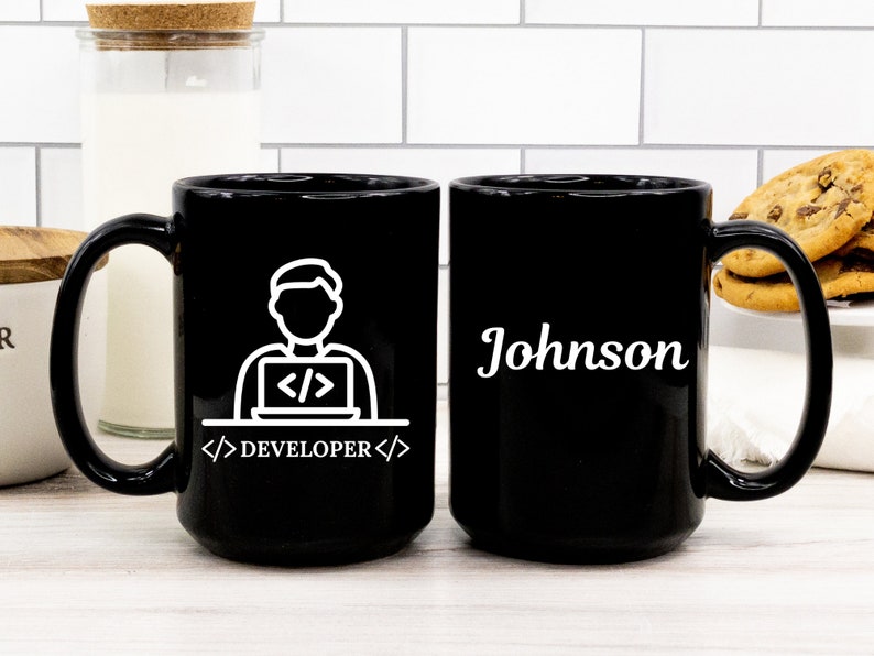 May include: Black ceramic mug with a white graphic of a person working on a laptop, with the word "DEVELOPER" below. The other side of the mug has the name "Johnson" in white. The mug is on a white surface.