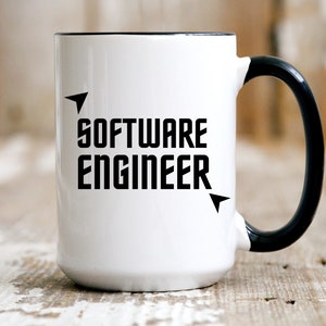 Puede incluir: Taza de cer&aacute;mica blanca con asa y borde negros. La taza presenta las palabras "SOFTWARE ENGINEER" en negrita, letras may&uacute;sculas negras, con elementos de dise&ntilde;o en forma de flecha. La taza est&aacute; sobre una superficie de madera r&uacute;stica.