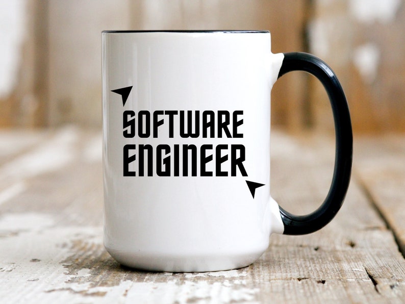 Puede incluir: Taza de cer&aacute;mica blanca con asa y borde negros. La taza presenta las palabras "SOFTWARE ENGINEER" en negrita, letras may&uacute;sculas negras, con elementos de dise&ntilde;o en forma de flecha. La taza est&aacute; sobre una superficie de madera r&uacute;stica.