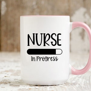 May include: White ceramic coffee mug with a pink rim and handle. The mug has black text that reads "NURSE" with a progress bar underneath and the words "In Progress".