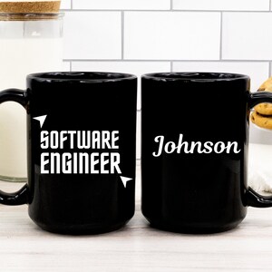 Puede incluir: Dos tazas de cer&aacute;mica negra. Una taza tiene el texto "SOFTWARE ENGINEER" en blanco. La otra taza tiene el nombre "Johnson" en blanco. Ambas tazas tienen un asa grande y est&aacute;n sobre una superficie blanca.