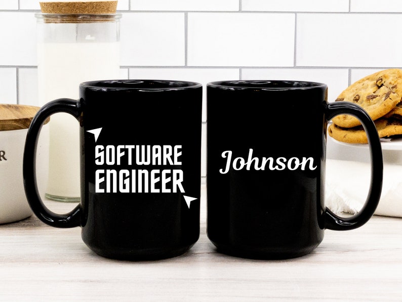 Puede incluir: Dos tazas de cer&aacute;mica negra. Una taza tiene el texto "SOFTWARE ENGINEER" en blanco. La otra taza tiene el nombre "Johnson" en blanco. Ambas tazas tienen un asa grande y est&aacute;n sobre una superficie blanca.