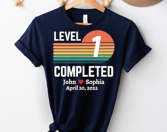 May include: Navy blue t-shirt with a retro-style graphic that says "Level 1 Completed" with a large number 1 in a circle. The graphic also includes a rainbow stripe design. Below the graphic, the text reads "John ♥ Sophia April 20, 2022".