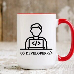 May include: White ceramic mug with a red interior and handle. The mug features a black line drawing of a person working on a laptop, with the word "DEVELOPER" below. The design is simple and modern, suitable for a tech professional.