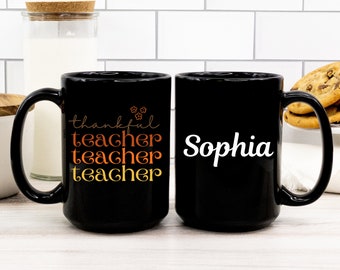 Może przedstawiać: Dwa czarne ceramiczne kubki. Jeden kubek ma napis "thankful teacher teacher teacher" w kolorze pomarańczowym i żółtym. Drugi kubek ma imię "Sophia" w kolorze białym. Kubki stoją na białej powierzchni.