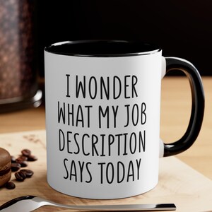 May include: White ceramic mug with a black interior and handle. The mug features the text "I WONDER WHAT MY JOB DESCRIPTION SAYS TODAY" in black, bold letters. The mug is on a wooden surface.