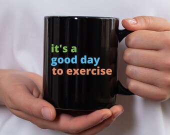 Może przedstawiać: Czarny ceramiczny kubek z napisem "it's a good day to exercise" w kolorze zielonym, niebieskim i pomarańczowym. Kubek jest trzymany przez osobę.