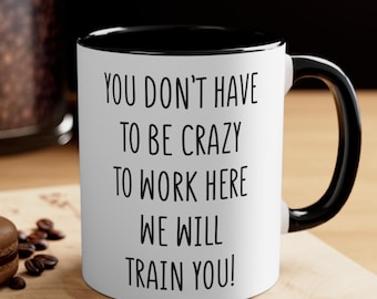May include: White ceramic mug with a black interior and handle. The mug features the text "YOU DON'T HAVE TO BE CRAZY TO WORK HERE WE WILL TRAIN YOU!" in black, bold font. The mug is on a wooden surface with coffee beans and a spoon.