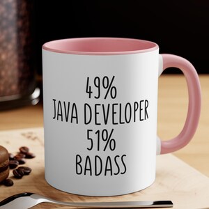 May include: White ceramic mug with a pink interior and handle. The mug features the text "49% JAVA DEVELOPER 51% BADASS" in black, bold font. The mug is sitting on a wooden surface.