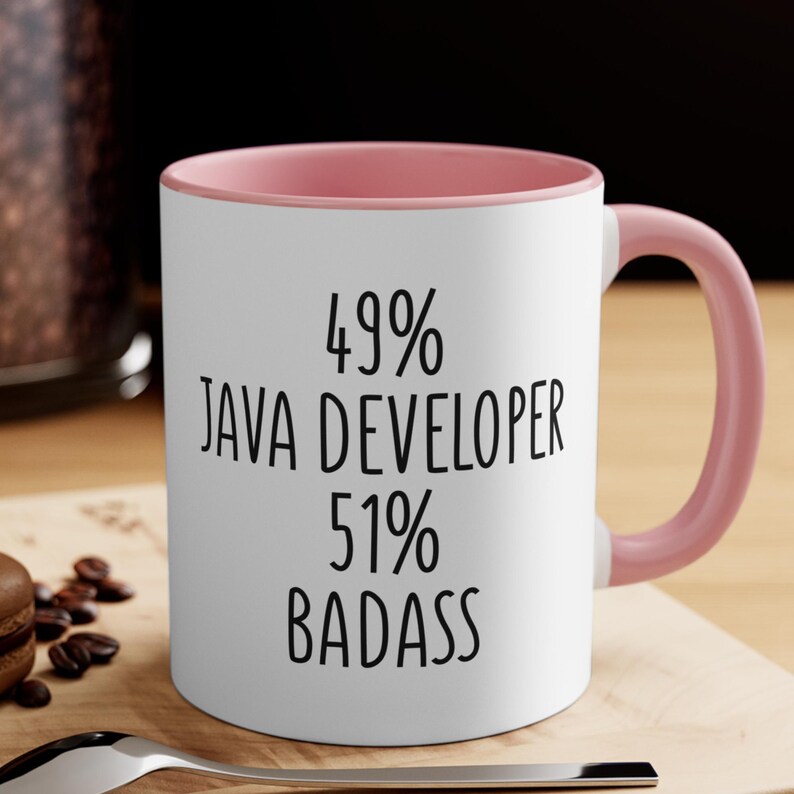 May include: White ceramic mug with a pink interior and handle. The mug features the text "49% JAVA DEVELOPER 51% BADASS" in black, bold font. The mug is sitting on a wooden surface.