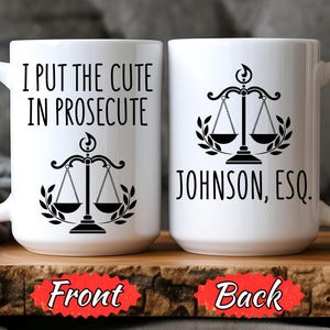 May include: White ceramic mug with the text "I PUT THE CUTE IN PROSECUTE" and a scales of justice graphic. The back of the mug has the name "JOHNSON, ESQ." and the same graphic. The mug is on a wooden surface.