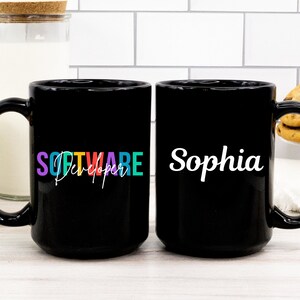 May include: Two black coffee mugs. One mug has the word "SOFTWARE" in purple, orange, and teal, with "Developer" in white script. The other mug has the name "Sophia" in white script. The mugs are on a white surface.