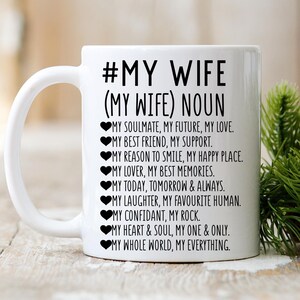 以下が含まれることがあります： 「#MY WIFE (MY WIFE) NOUN」のテキストと愛情あふれる説明が書かれた白いセラミックマグ。デザイン全体に黒い文字とハートのシンボルが使用されています。マグカップには曲がったハンドルがあり、背景はぼやけています。