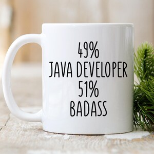 May include: White ceramic mug with the text "49% JAVA DEVELOPER 51% BADASS" in black. The mug has a curved handle and is set against a blurred background with greenery.