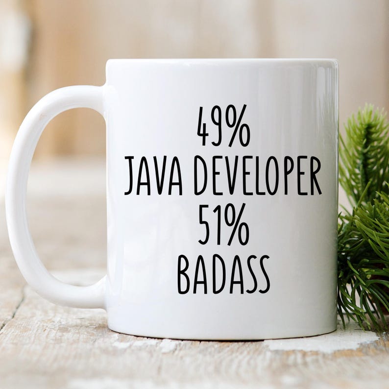May include: White ceramic mug with the text "49% JAVA DEVELOPER 51% BADASS" in black. The mug has a curved handle and is set against a blurred background with greenery.