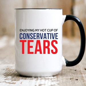 May include: White ceramic coffee mug with a black rim and handle. The mug has the text "Enjoying my hot cup of Conservative Tears" printed on it in blue and red.