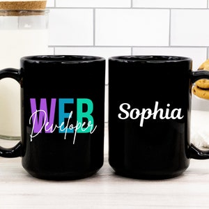 Peut inclure: Deux mugs en céramique noirs. L'un porte les mots "WEB Developer" en violet, bleu sarcelle et vert, avec une police blanche manuscrite. L'autre mug porte le nom "Sophia" en blanc. Les mugs sont sur une surface blanche.