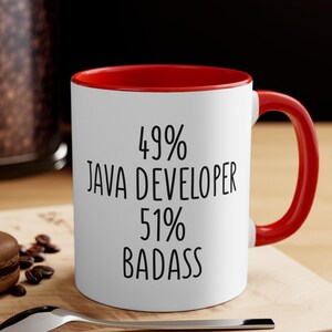 May include: White ceramic mug with a red interior and handle. The mug features the text "49% JAVA DEVELOPER 51% BADASS" in black, bold font. The mug is sitting on a wooden surface.