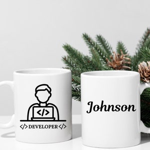 May include: Two white ceramic mugs. One mug features a black line drawing of a person working on a laptop with the text "DEVELOPER". The other mug has the name "Johnson" in a cursive font.
