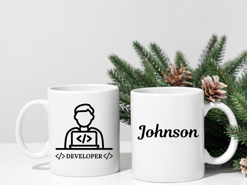 May include: Two white ceramic mugs. One mug features a black line drawing of a person working on a laptop with the text "DEVELOPER". The other mug has the name "Johnson" in a cursive font.