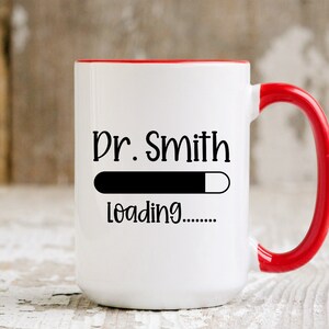 May include: White ceramic mug with a red interior and handle. The mug features the text "Dr. Smith" above a loading bar graphic, with "loading......" below. The mug is designed for beverages.