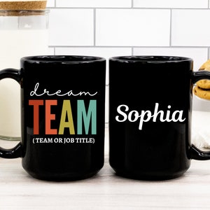 May include: Two black coffee mugs. One mug has the text "dream TEAM (TEAM OR JOB TITLE)" in white with the word "TEAM" in rainbow colors. The other mug has the name "Sophia" in white cursive.