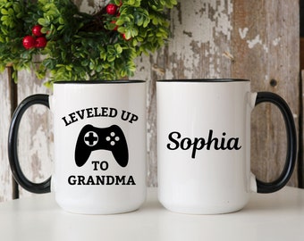 May include: White ceramic mug with a black handle and rim. One mug features the text "LEVELED UP TO GRANDMA" with a black video game controller graphic. The other mug has the name "Sophia" in black script.