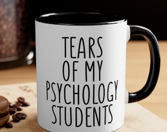 May include: White ceramic mug with a black interior and handle. The mug features the text "TEARS OF MY PSYCHOLOGY STUDENTS" in black, centered on the front. The mug is on a wooden surface with a spoon and coffee beans.