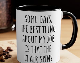 May include: White ceramic mug with a black interior and handle. The mug features the text "SOME DAYS, THE BEST THING ABOUT MY JOB IS THAT THE CHAIR SPINS" in black lettering. The mug is on a wooden surface.