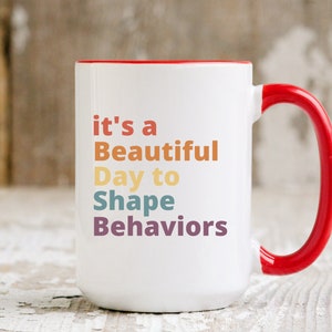 May include: White ceramic mug with a red rim and handle. The mug has the text "it's a Beautiful Day to Shape Behaviors" printed on it in a rainbow of colors.