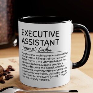 May include: White ceramic mug with a black interior and handle. The mug features the text "EXECUTIVE ASSISTANT" in large black letters, with a definition and the name "Sophia" below. The mug is on a wooden surface.