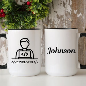 May include: Two white ceramic mugs with black handles and rims. One mug features a black and white graphic of a person coding on a laptop with the text "DEVELOPER". The other mug has the name "Johnson" in black script.