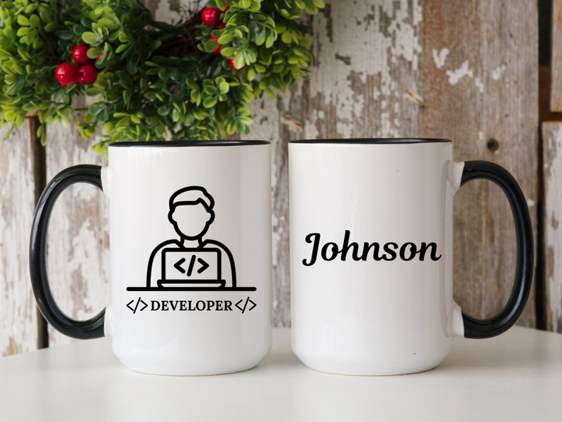 May include: Two white ceramic mugs with black handles and rims. One mug features a black and white graphic of a person coding on a laptop with the text "DEVELOPER". The other mug has the name "Johnson" in black script.