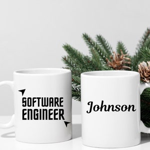Puede incluir: Dos tazas de cer&aacute;mica blancas. Una taza tiene el texto "SOFTWARE ENGINEER" en negrita negra con detalles de flechas. La otra taza tiene el nombre "Johnson" en cursiva negra. Ramas de pino verdes y pi&ntilde;as en el fondo.