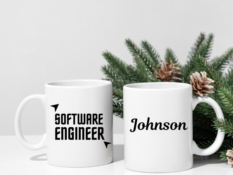 Puede incluir: Dos tazas de cer&aacute;mica blancas. Una taza tiene el texto "SOFTWARE ENGINEER" en negrita negra con detalles de flechas. La otra taza tiene el nombre "Johnson" en cursiva negra. Ramas de pino verdes y pi&ntilde;as en el fondo.
